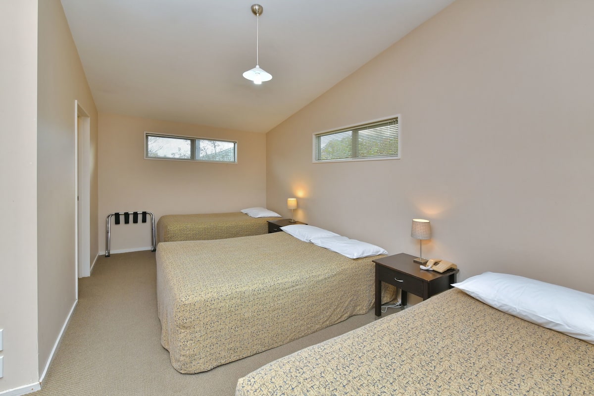Quarto de hotel com duas camas, um radiador, uma mesa com luminária e duas janelas.