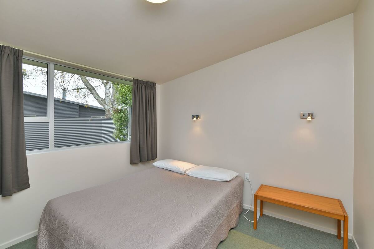 Quarto com uma cama de solteiro, um banco, duas luzes fixadas na parede e uma janela com cortinas.