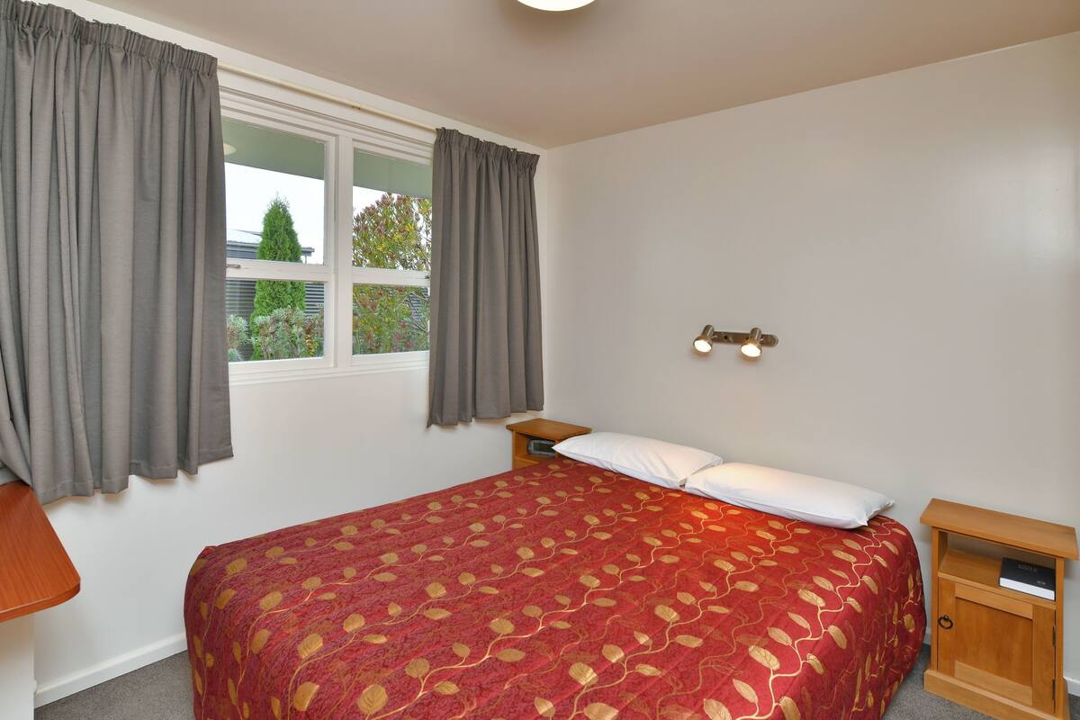 Quarto com cama, mesa de cabeceira, cortinas na janela e luminária na parede.