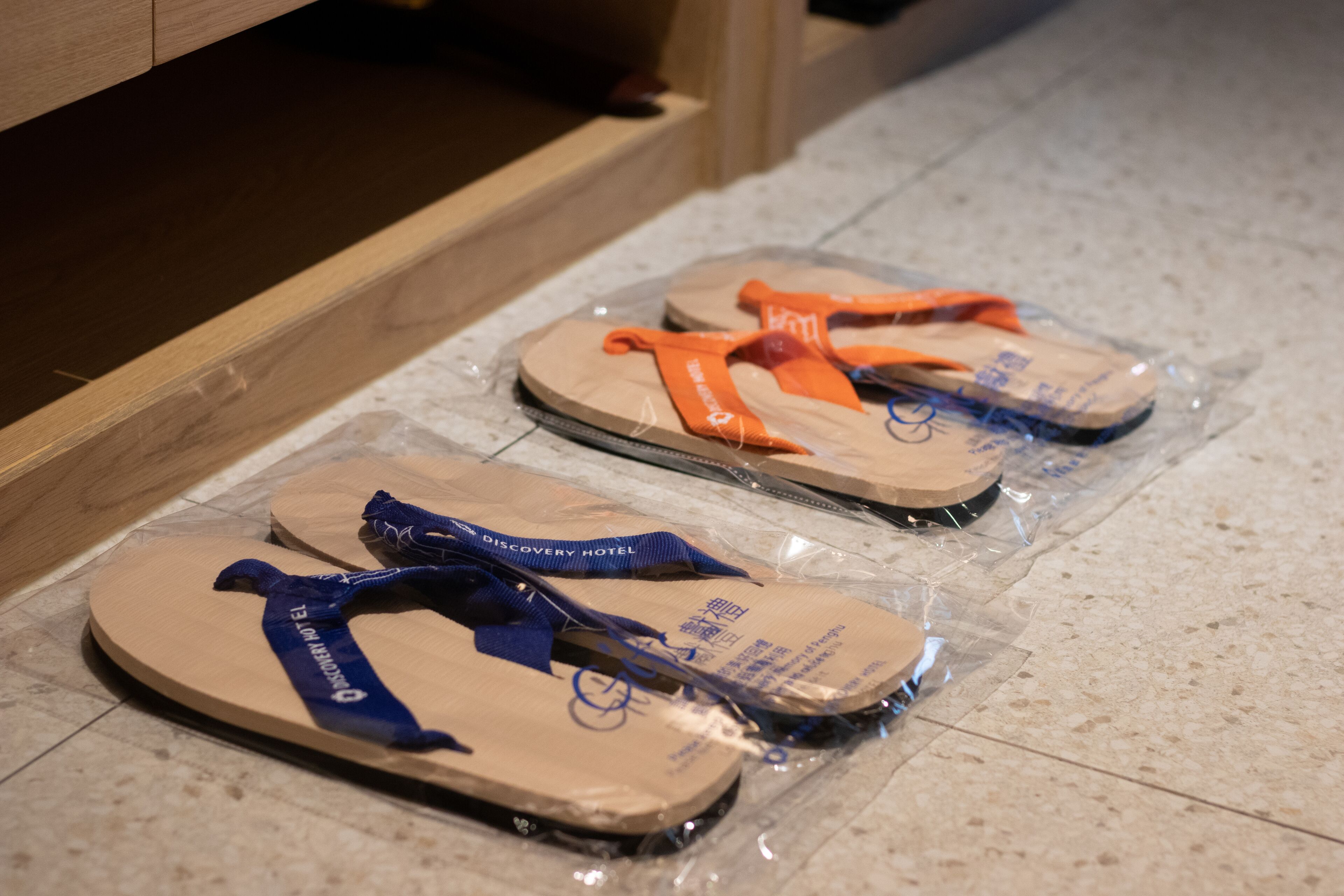 Une paire de tongs, une avec une bride bleue et l’autre avec une bride orange, toutes deux emballées dans du plastique, posées sur un sol carrelé.