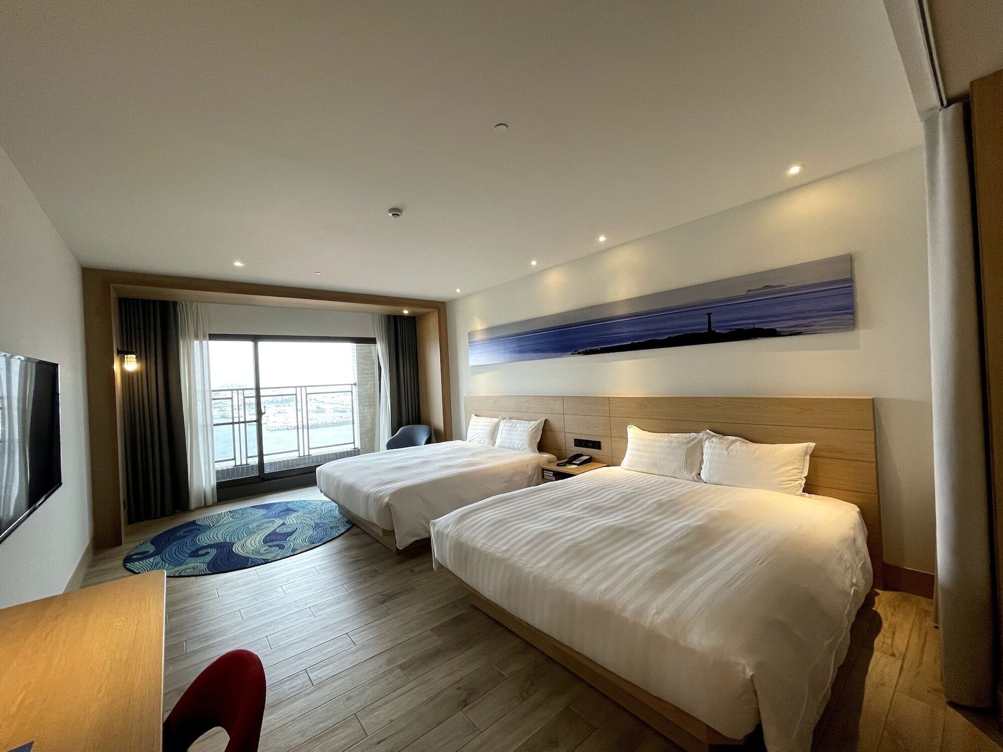 A hotel room with two beds, a TV, a desk, and a large window with a view.