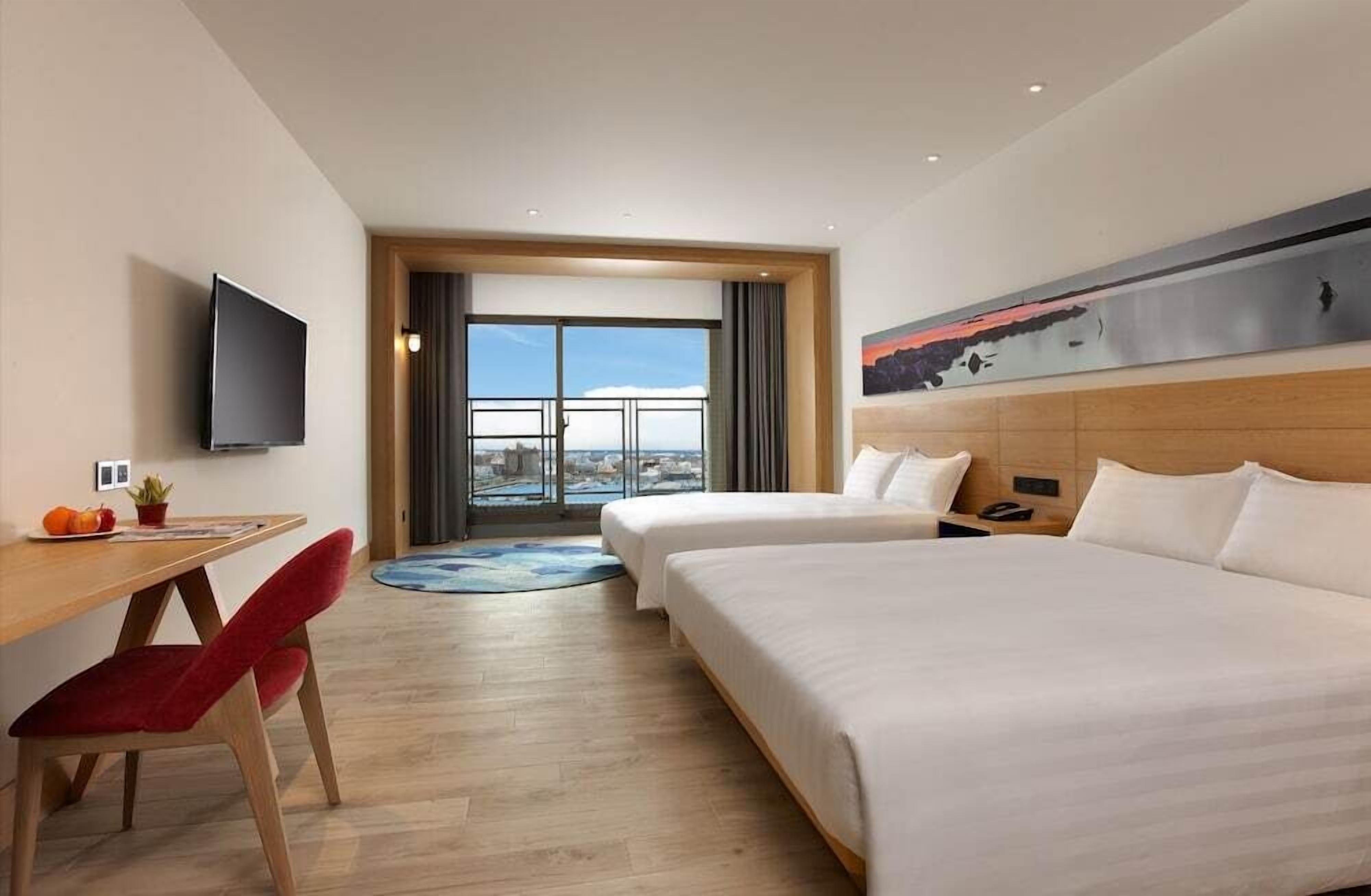 Une chambre d’hôtel équipée d’un lit, d’un bureau, d’une chaise, d’une télévision et d’une grande fenêtre donnant sur la ville.