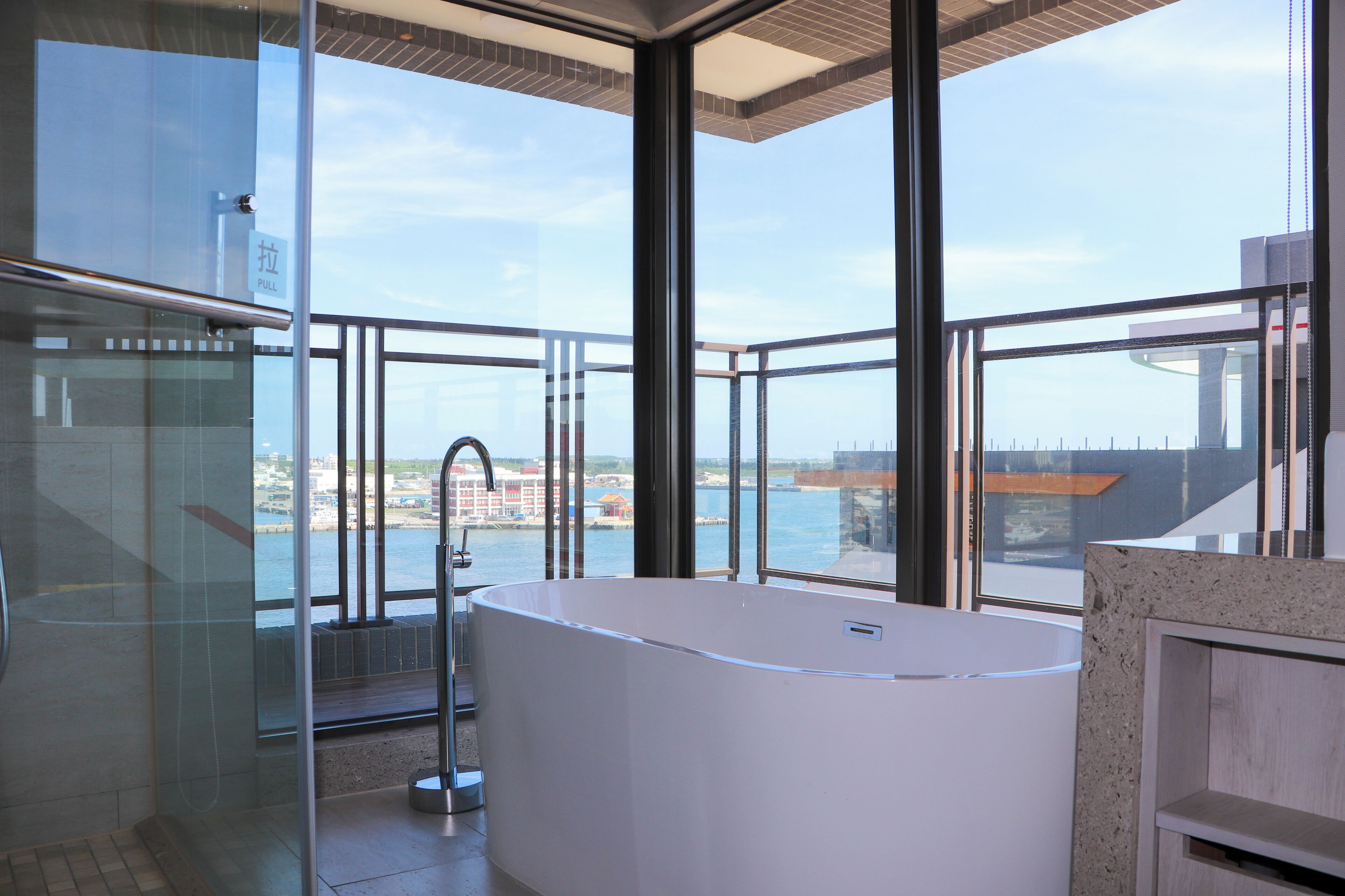 A modern bathroom with a large bathtub, glass shower enclosure, and a view of the sea and buildings.