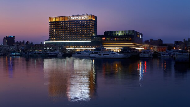 Ein modernes Hotel mit einem gut sichtbaren Schild "Discovery Hotel", einer Lage am Wasser und mehreren Booten, die im Yachthafen vor Anker liegen.