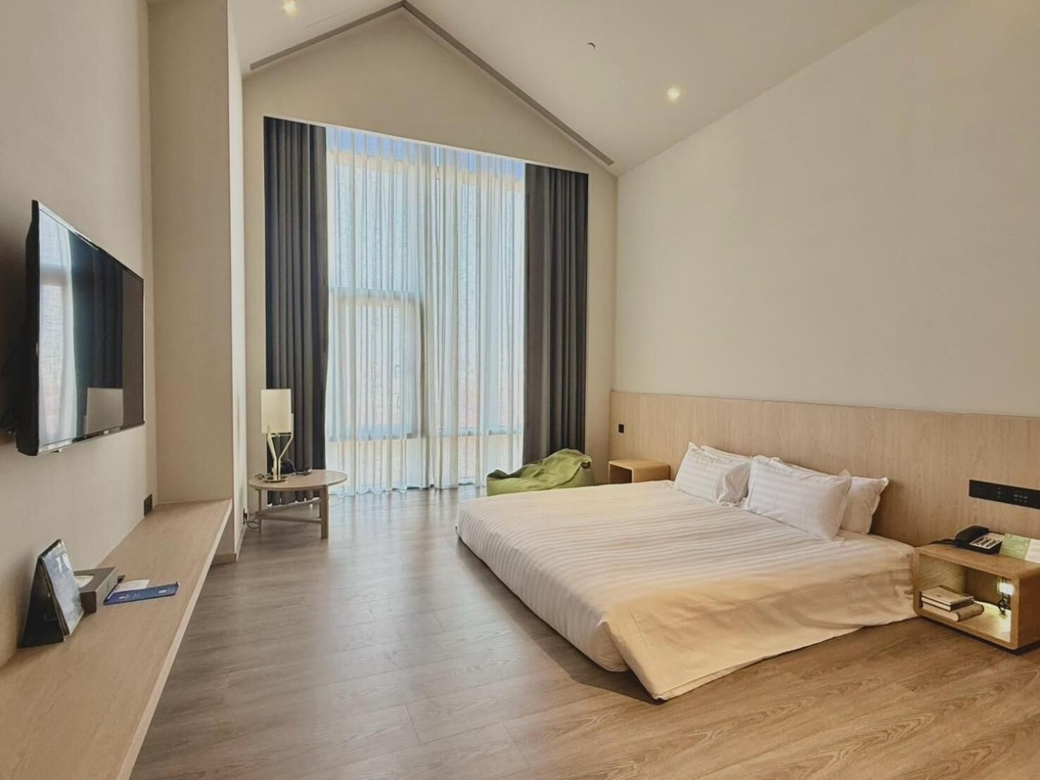 Una habitación de hotel moderna con una cama grande, un televisor de pantalla plana, un escritorio con lámpara y vistas a la ciudad a través de amplios ventanales.