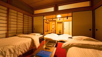 A traditional Japanese-style hotel room with tatami flooring, a low wooden ceiling, and sliding shoji doors. There are two beds with white bedding and a small table with a blue book and a remote control.