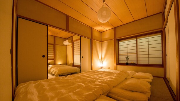 Ein traditioneller japanischer Raum mit Tatami-Bodenbelag, einem niedrigen Bett mit weißer Bettwäsche, Schiebetüren im Shoji-Stil und einer hängenden Papierlaterne.