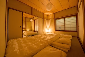 Une pièce de style japonais traditionnel, avec un sol recouvert de tatamis, un lit bas avec du linge de lit blanc, des portes coulissantes en papier de riz (shoji) et une lanterne en papier suspendue.