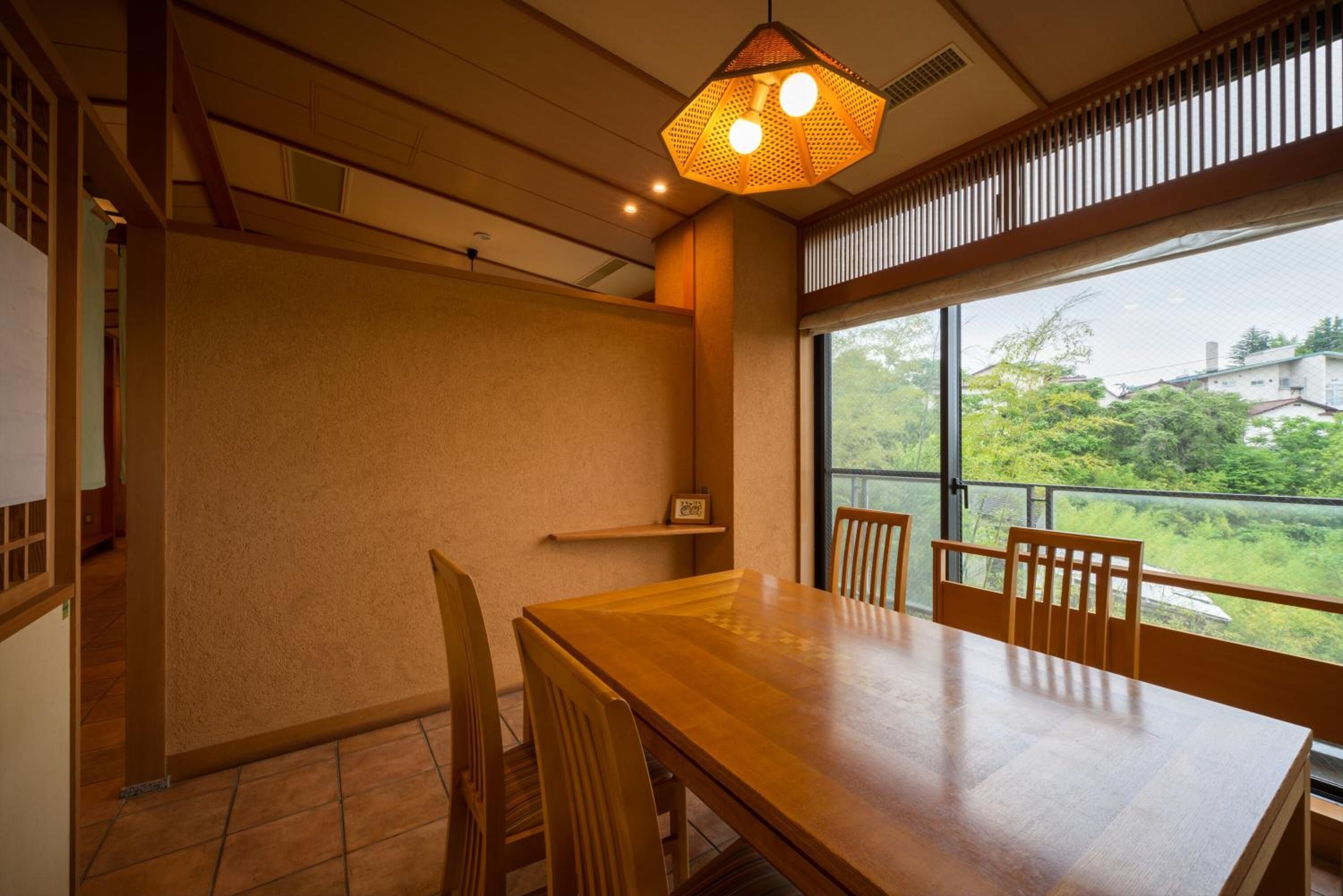 Un espace repas avec une table et des chaises en bois, un luminaire suspendu et une vue sur la verdure grâce à de grandes fenêtres.