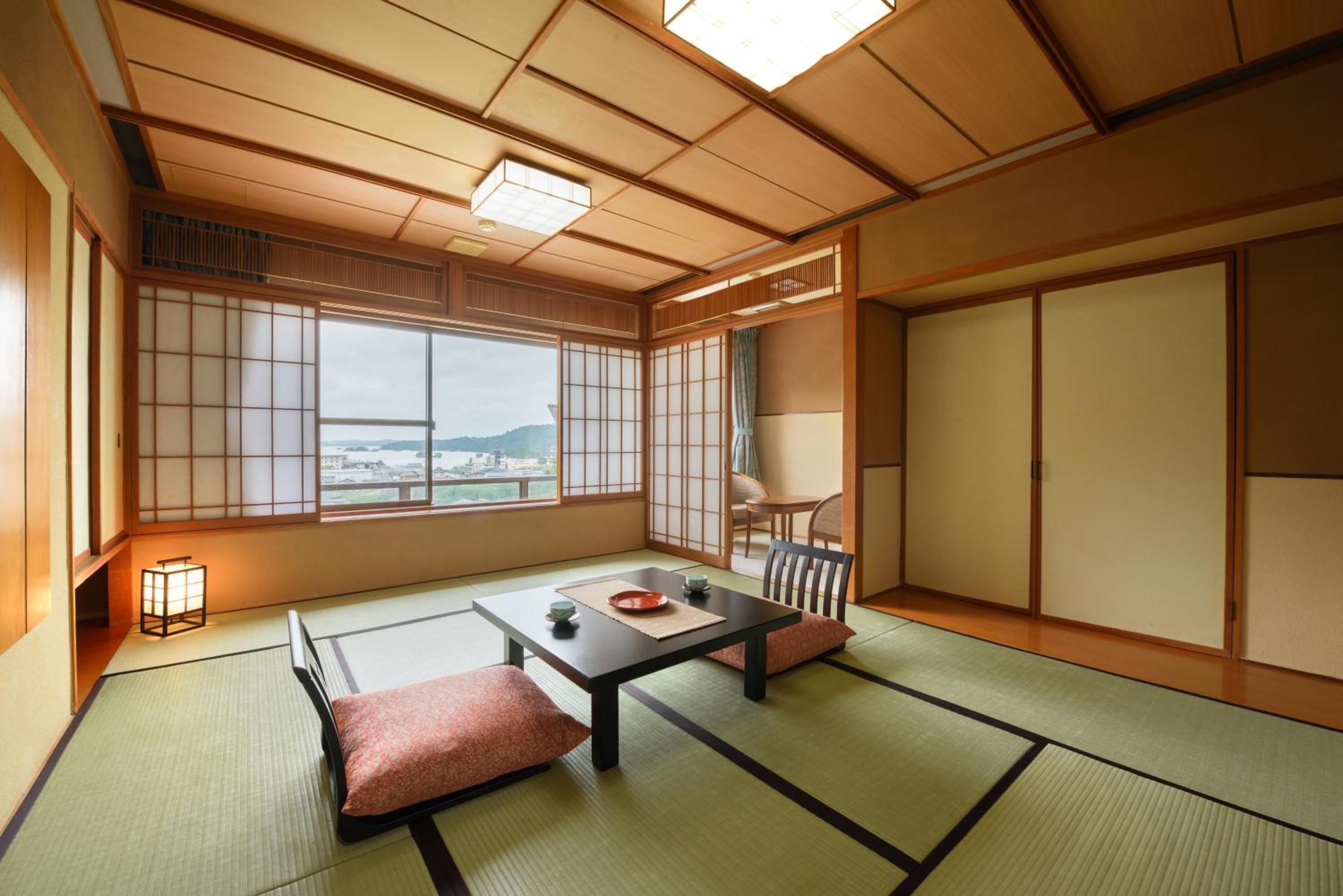 Ein traditioneller japanischer Raum mit Tatami-Böden, einem niedrigen Tisch mit Kissen, Schiebetüren und Blick auf eine Stadtlandschaft.