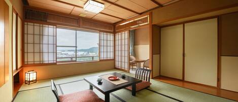 A traditional Japanese room with tatami flooring, a low table with cushions, sliding doors, and a view of a cityscape.