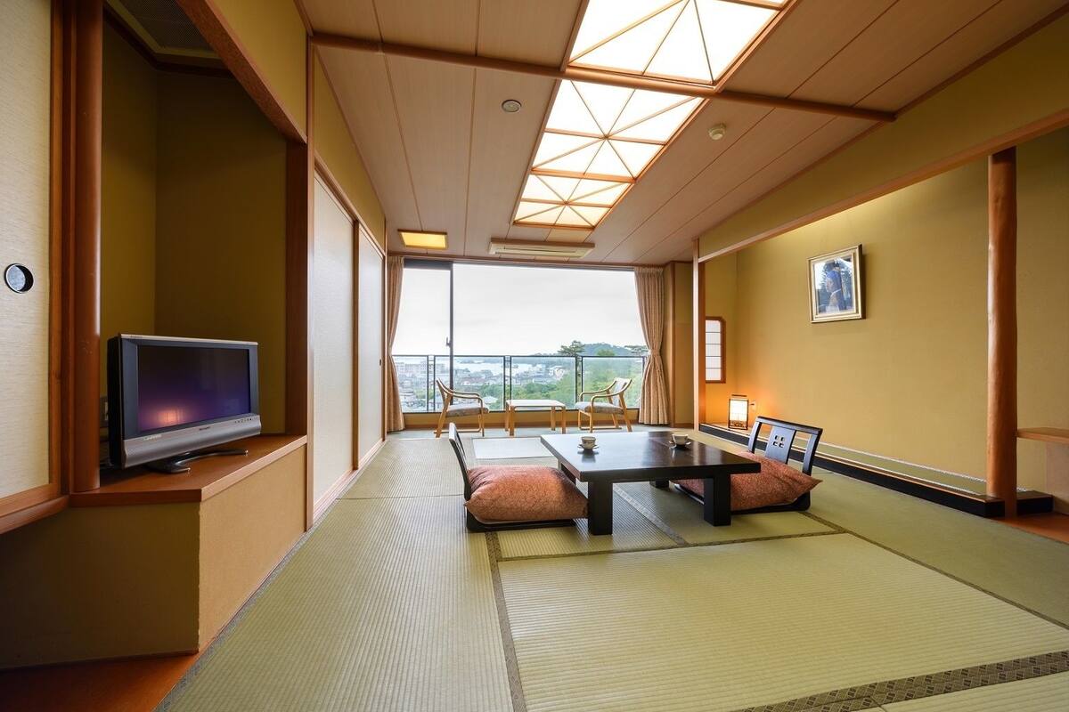 A traditional Japanese-style room with tatami flooring, a low table, cushions, a television, and a view of the city.