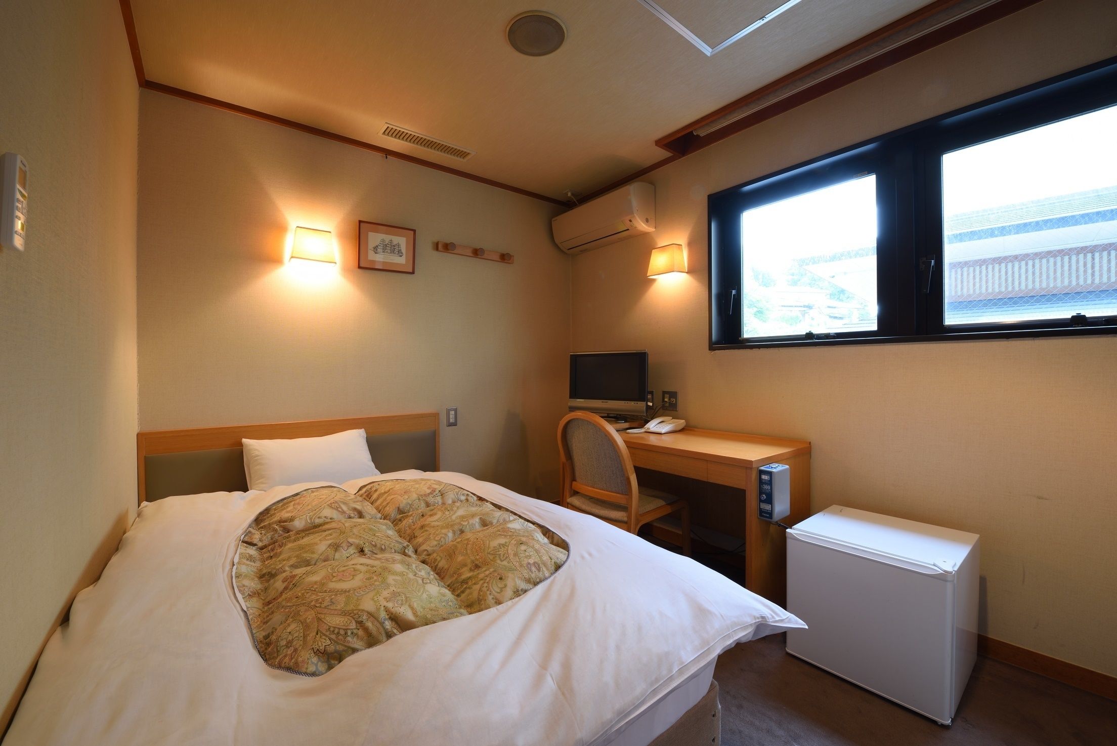 Habitación de hotel con cama, escritorio con computadora, refrigerador pequeño y ventana con vista a un edificio.