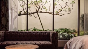 A room with a tufted sofa, a round wooden table, and a large window with a view of a tree branch and plants. - Green Palm (Yuanlin)
