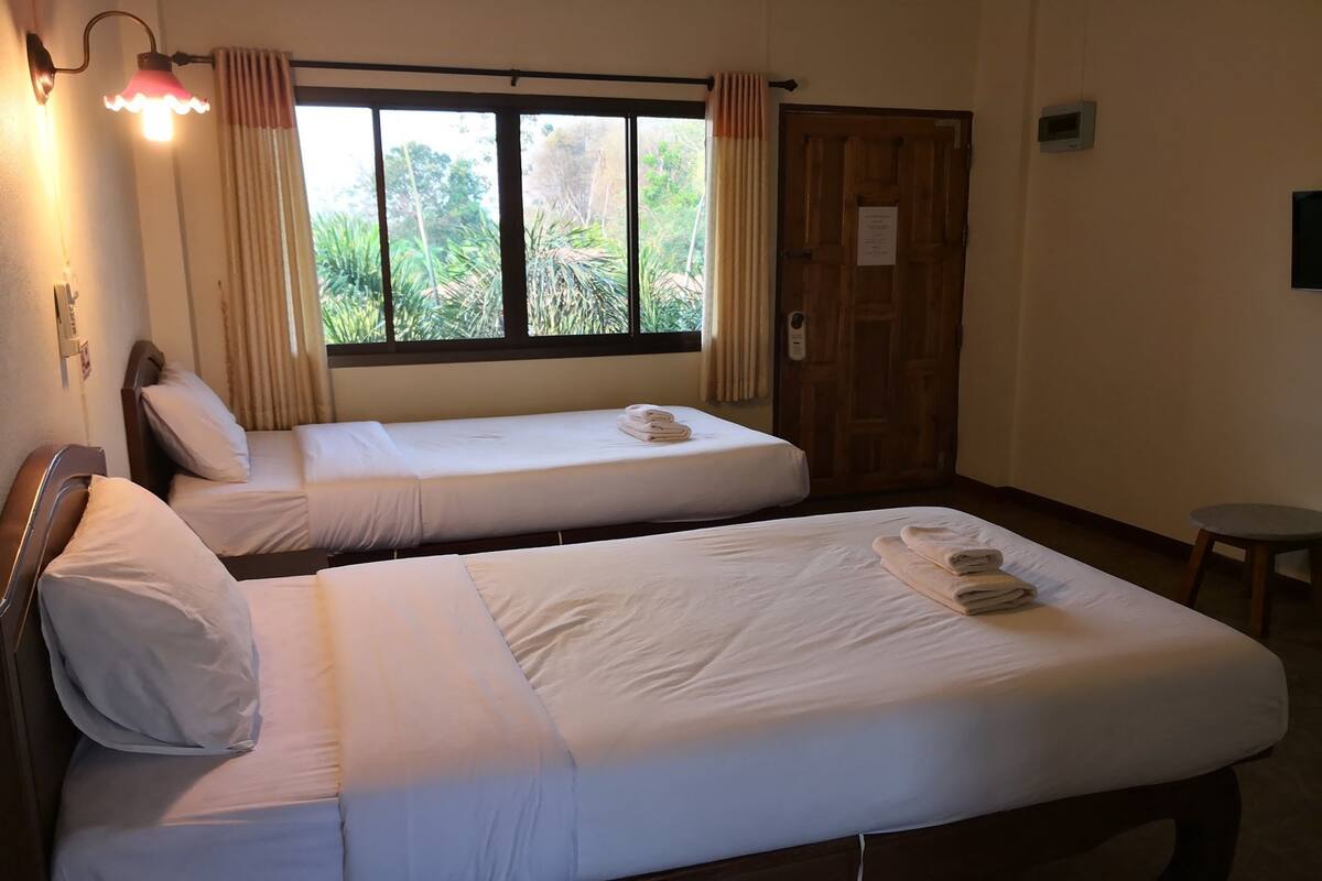 A hotel room with two single beds, a window with curtains, a wooden door, and a small stool.