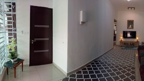 A hallway with a dark wooden door, a white wall, a patterned floor, a small table with a plant, and a sofa with cushions. - Khalish Homestay (Chemor)