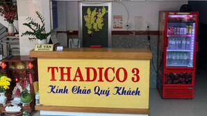 Ein Empfangstresen mit einem Schild, das "THADICO 3" und "Kinh Chào Quý Khách" zeigt. Darüber hängen Uhren, und an der Wand befinden sich Schilder mit den Aufschriften "NEW YORK", "VIET NAM" und "LONDON". Eine rote Getränkeautomaten steht rechts.