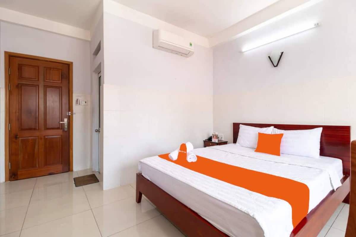Une chambre d’hôtel avec une porte en bois, un lit avec une literie blanche et orange, une table de chevet et un climatiseur fixé au mur.