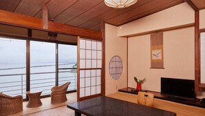 Een kamer met een groot raam met uitzicht op zee, een houten eettafel, rieten stoelen, een flatscreen televisie en een kleine potplant.