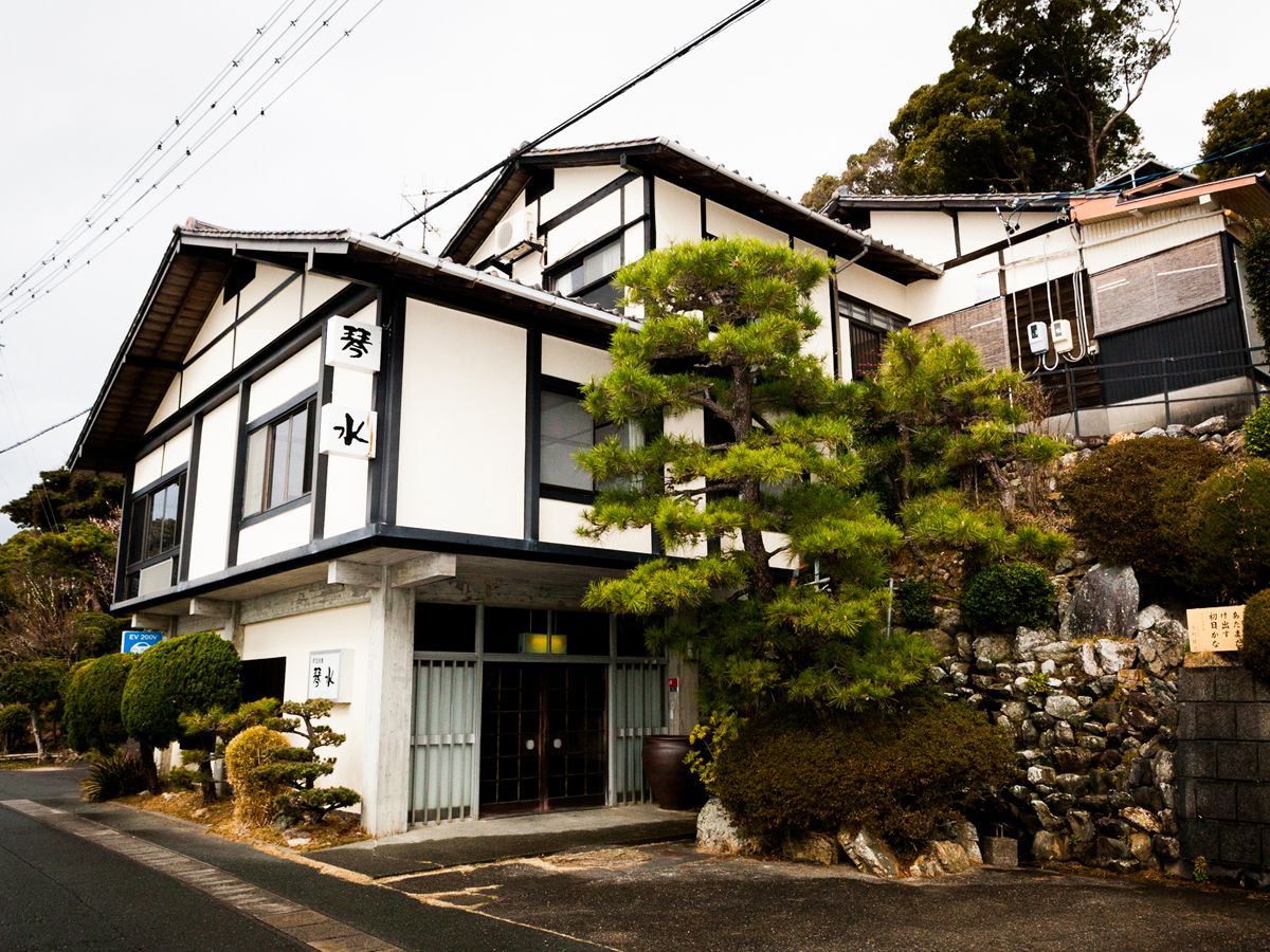A traditional Japanese building with a black and white exterior, a stone wall, and a well-maintained garden with various plants and a large tree.