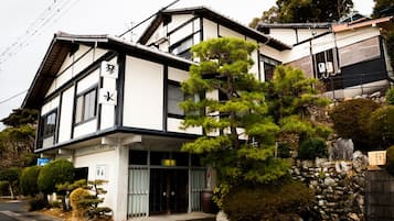 A traditional Japanese building with a black and white exterior, a stone wall, and a well-maintained garden with various plants and a large tree.