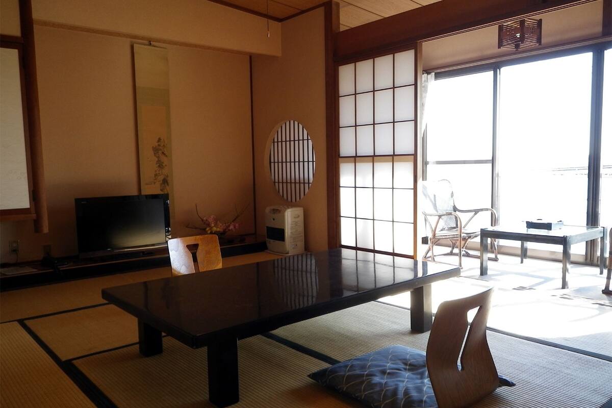 A traditional Japanese room with tatami flooring, a low table, a television, and a sliding door leading to a balcony.