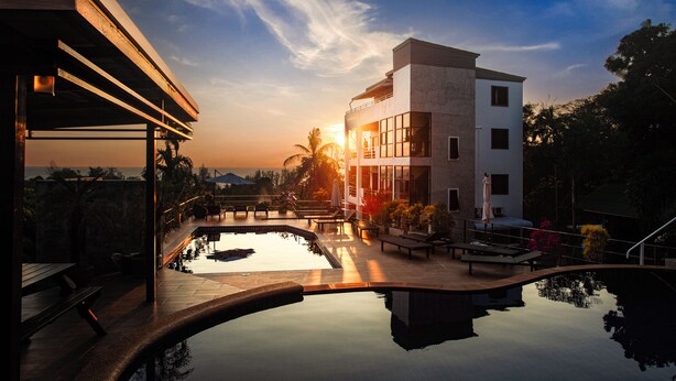 A modern hotel with a pool, lounge chairs, and a view of the sunset.