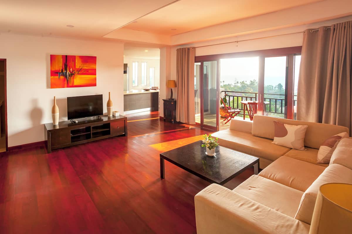 A spacious living room with a large sofa, a coffee table, and a flat-screen TV on a wooden console. A painting hangs on the wall, and there is a balcony with a view.