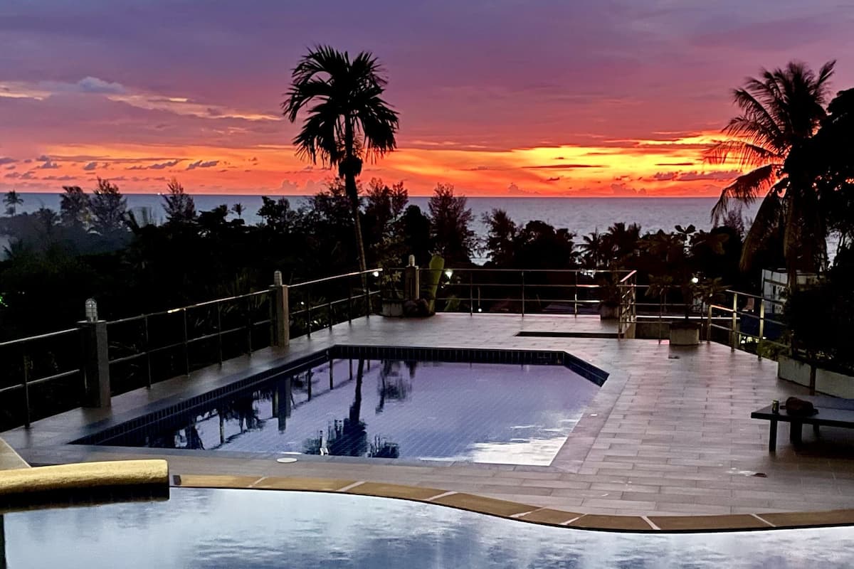 A swimming pool with a sunset view, surrounded by palm trees and a tiled deck.
