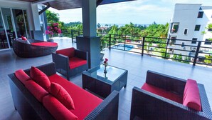 A covered outdoor seating area with wicker furniture and red cushions, overlooking a pool and lush greenery.