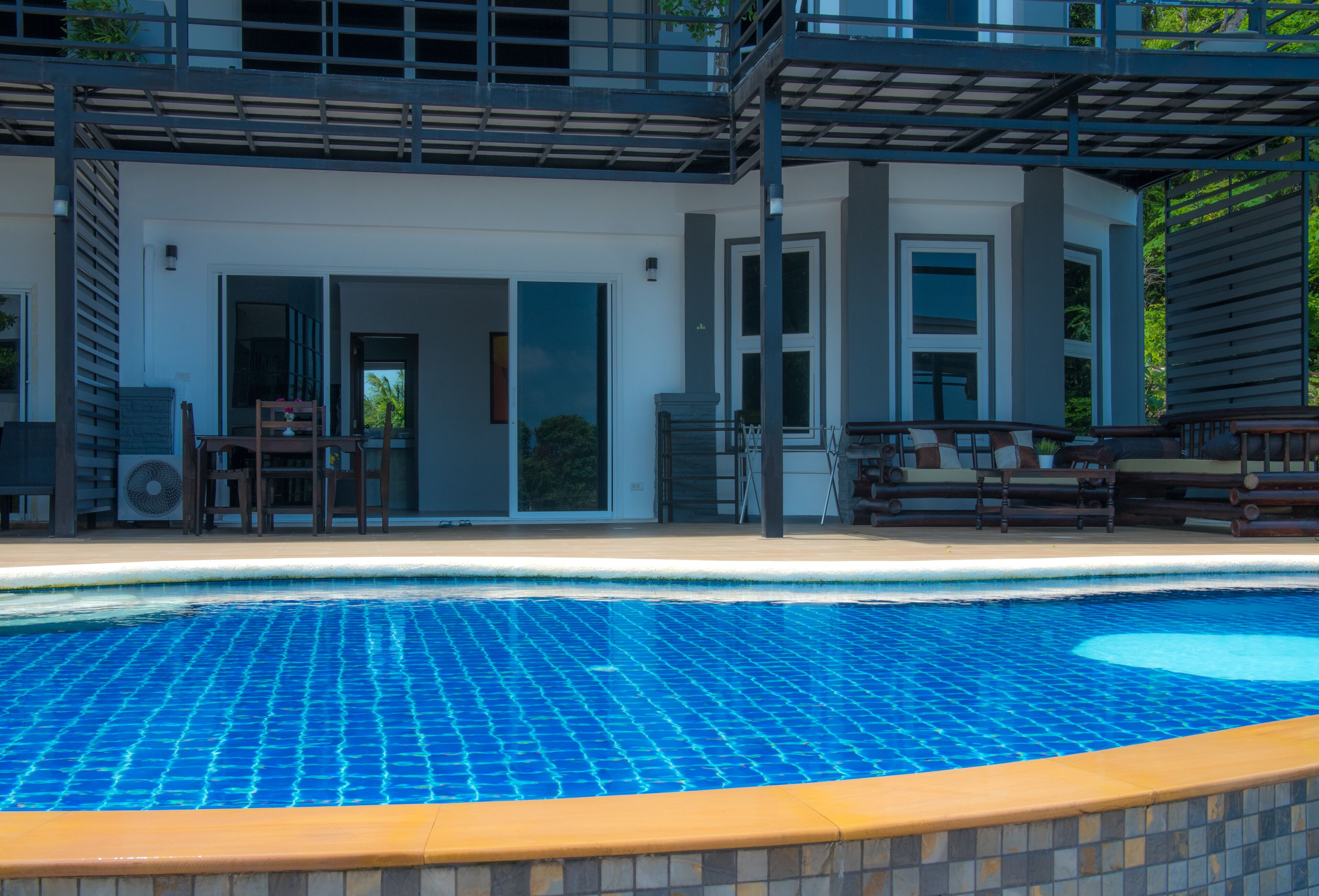 Un espace piscine moderne avec une piscine carrelée d’un bleu éclatant, un coin salon couvert avec des chaises, et un bâtiment doté de grandes fenêtres et d’un balcon.