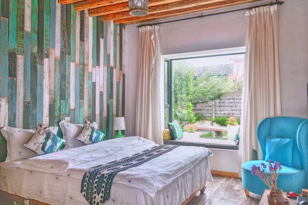 A bedroom with a bed, a blue armchair, a window with a view of greenery, and a wooden ceiling.