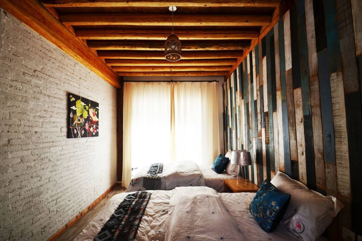 A bedroom with two beds, wooden beams, a hanging light fixture, and a window with curtains.
