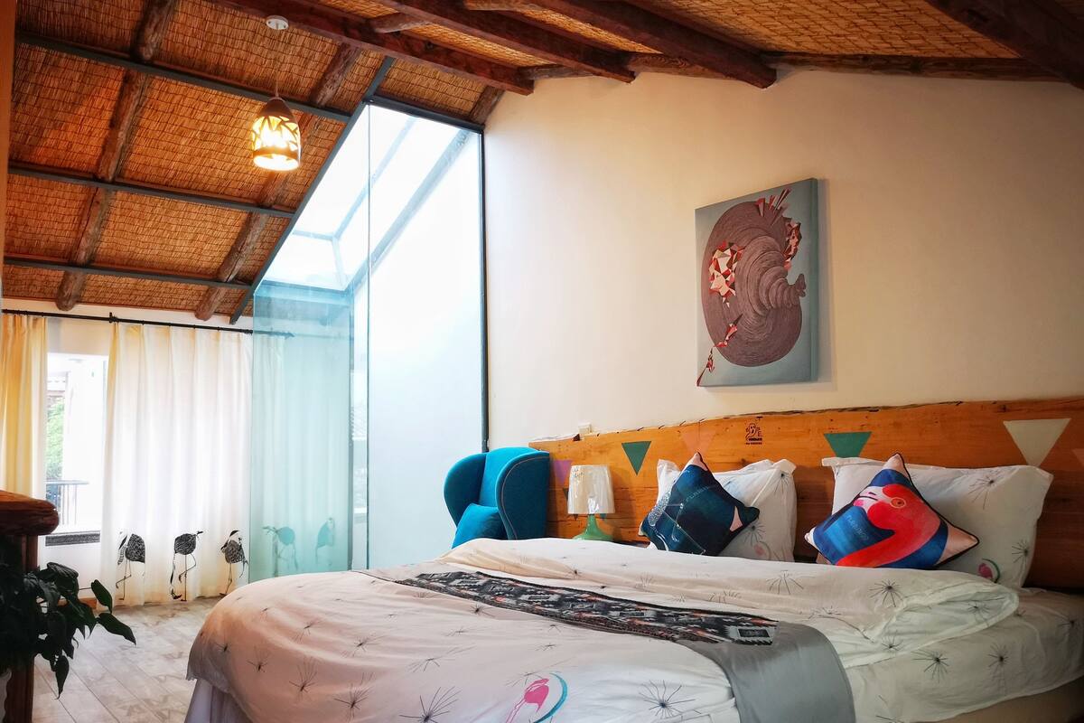 A hotel room with a large bed, a wooden headboard, a painting on the wall, and a window with curtains.
