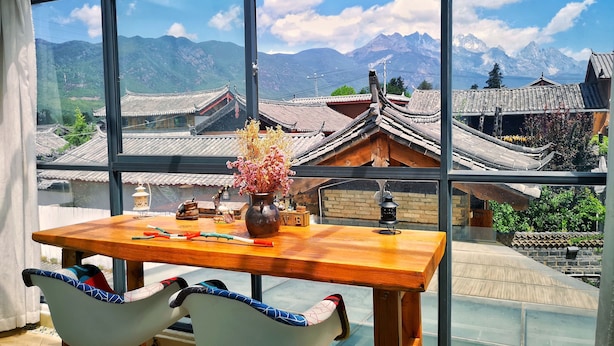 Uma sala de jantar com uma mesa e cadeiras, um vaso com flores e vista para edifícios tradicionais e montanhas através de grandes janelas.