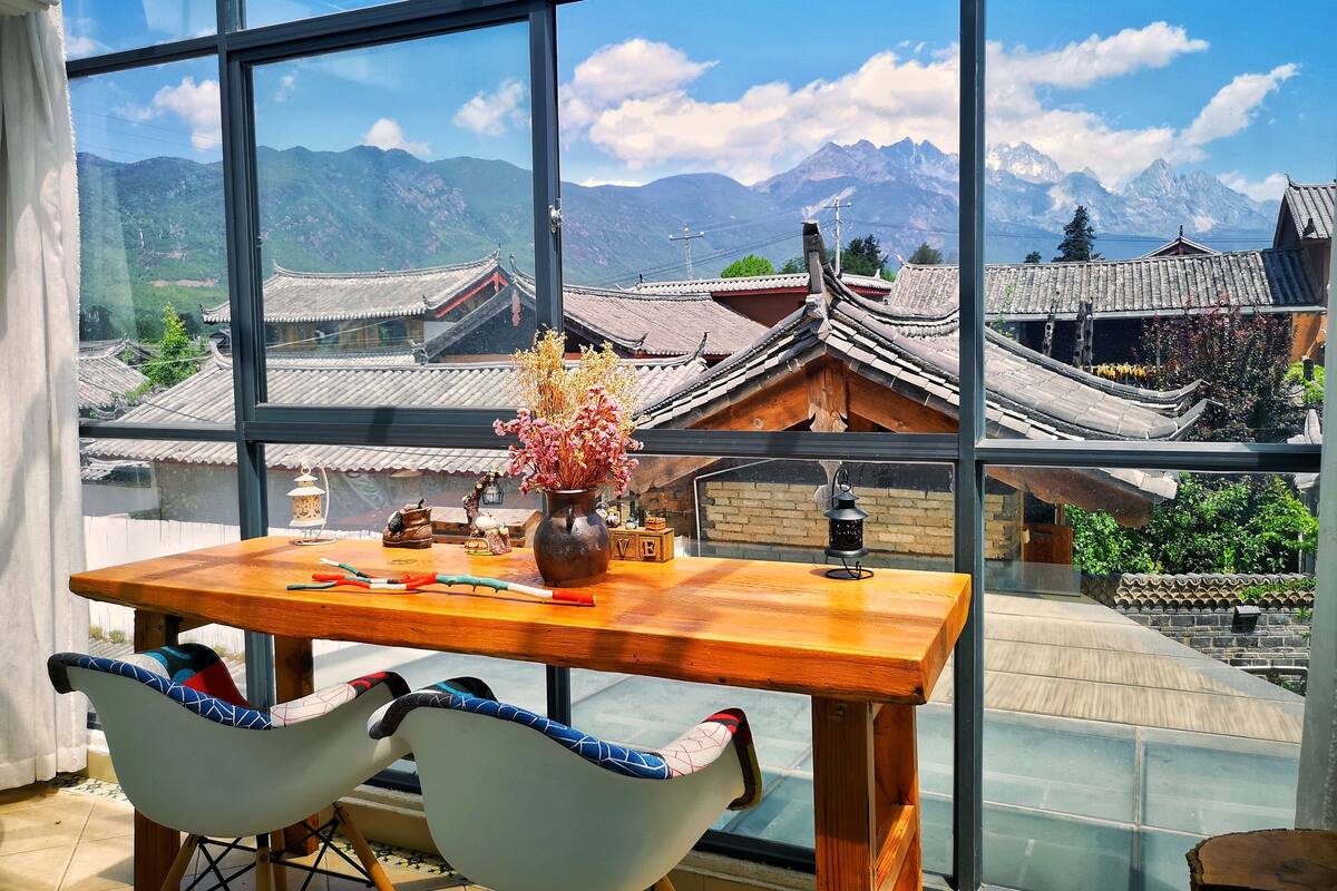 A dining area with a table and chairs, a vase with flowers, and a view of traditional buildings and mountains through large windows.