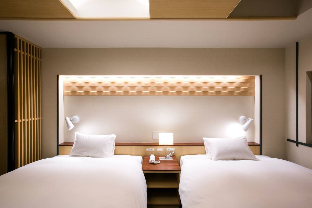 Habitación de hotel con dos camas, una mesita de noche con lámpara y un ventanal con persianas.