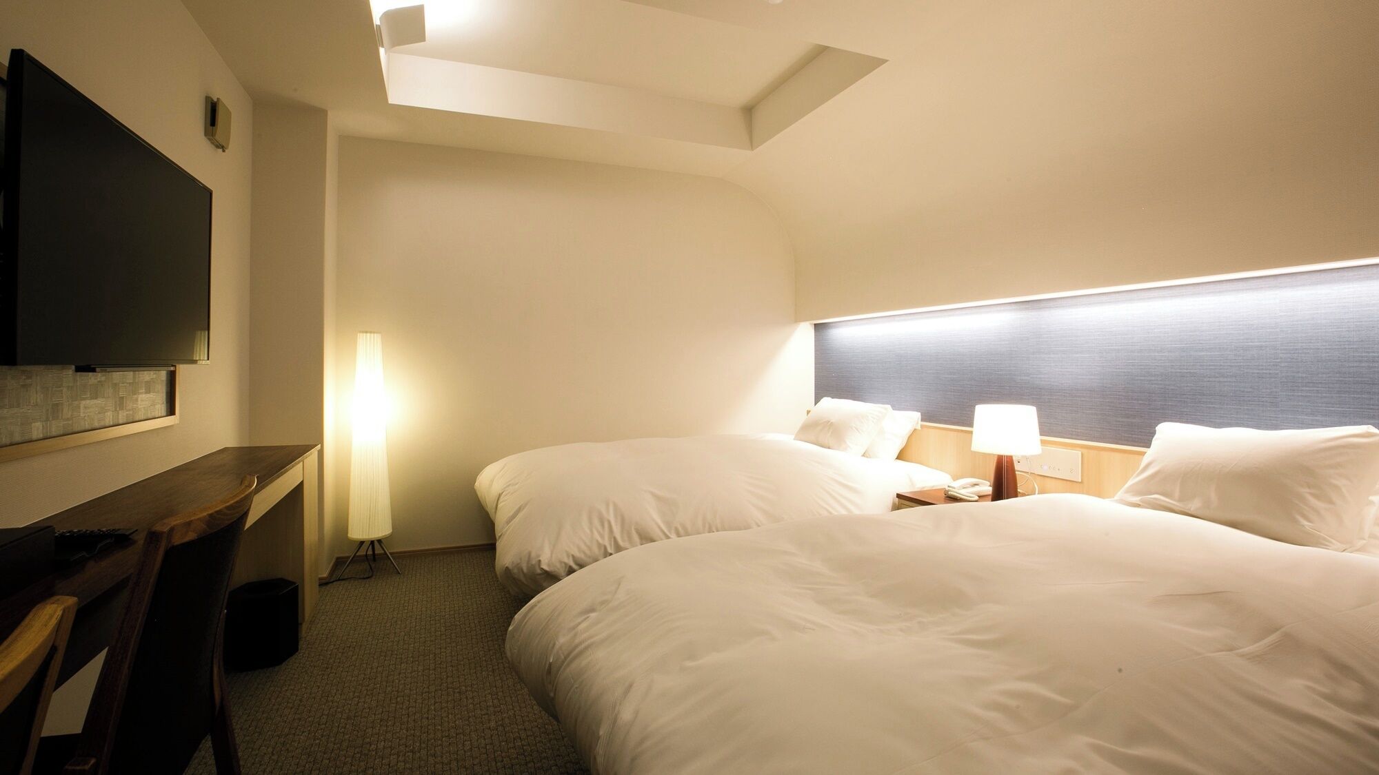 A hotel room with two beds, a desk, a TV, and a lamp.