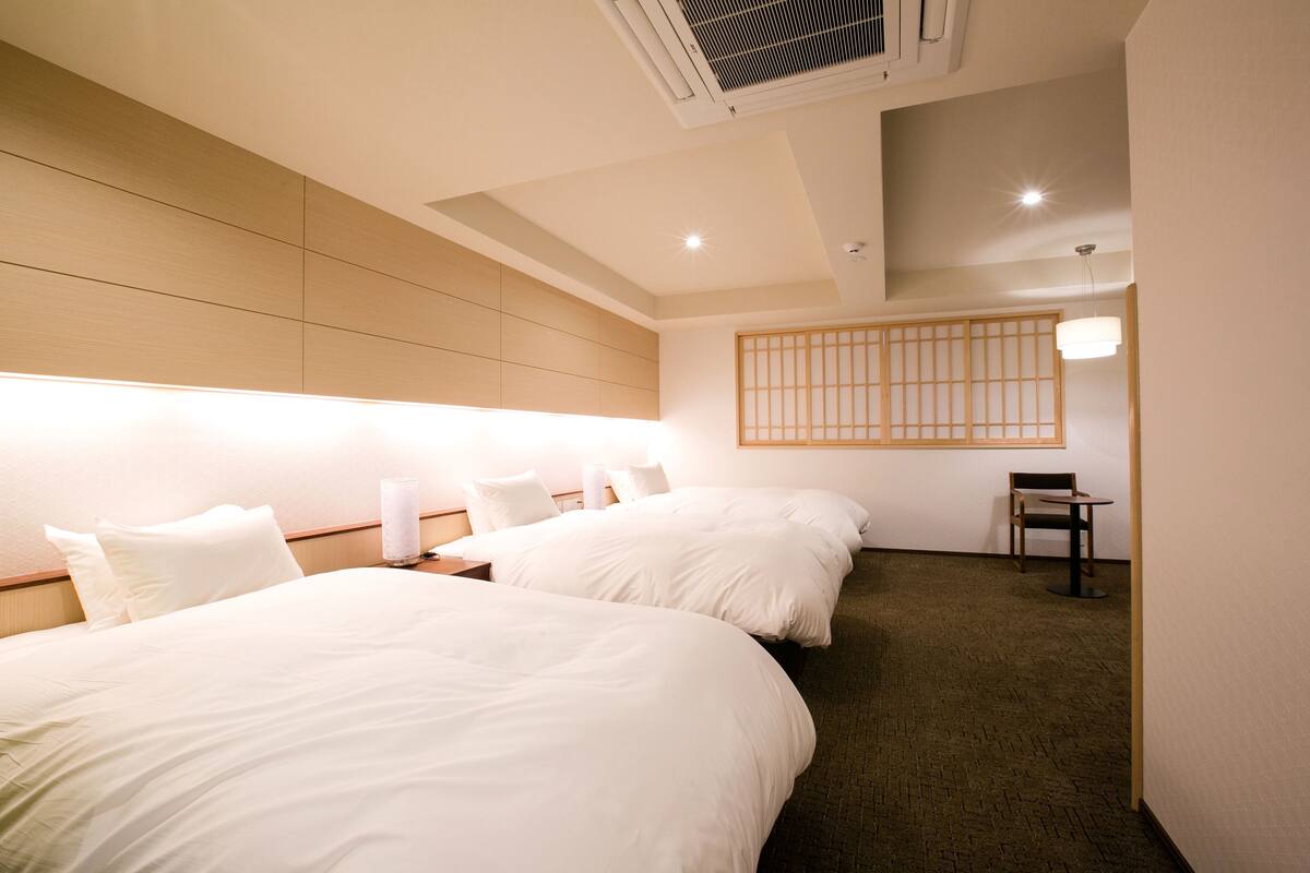 Habitación de hotel con dos camas, una mesita, una silla y un panel fijo en la pared.