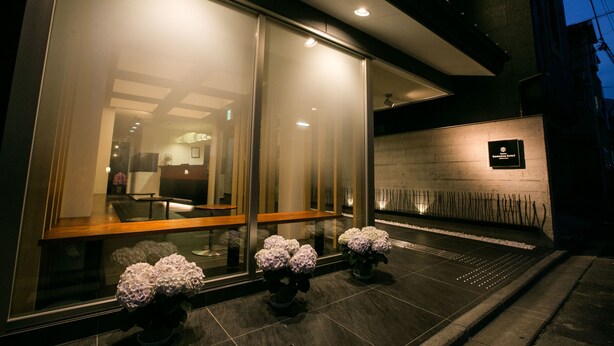 A modern hotel entrance with large glass doors, a reception desk, and potted plants.