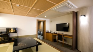A modern room with a tatami mat floor, a black table, a wooden desk, a television, and a kitchen area in the background.