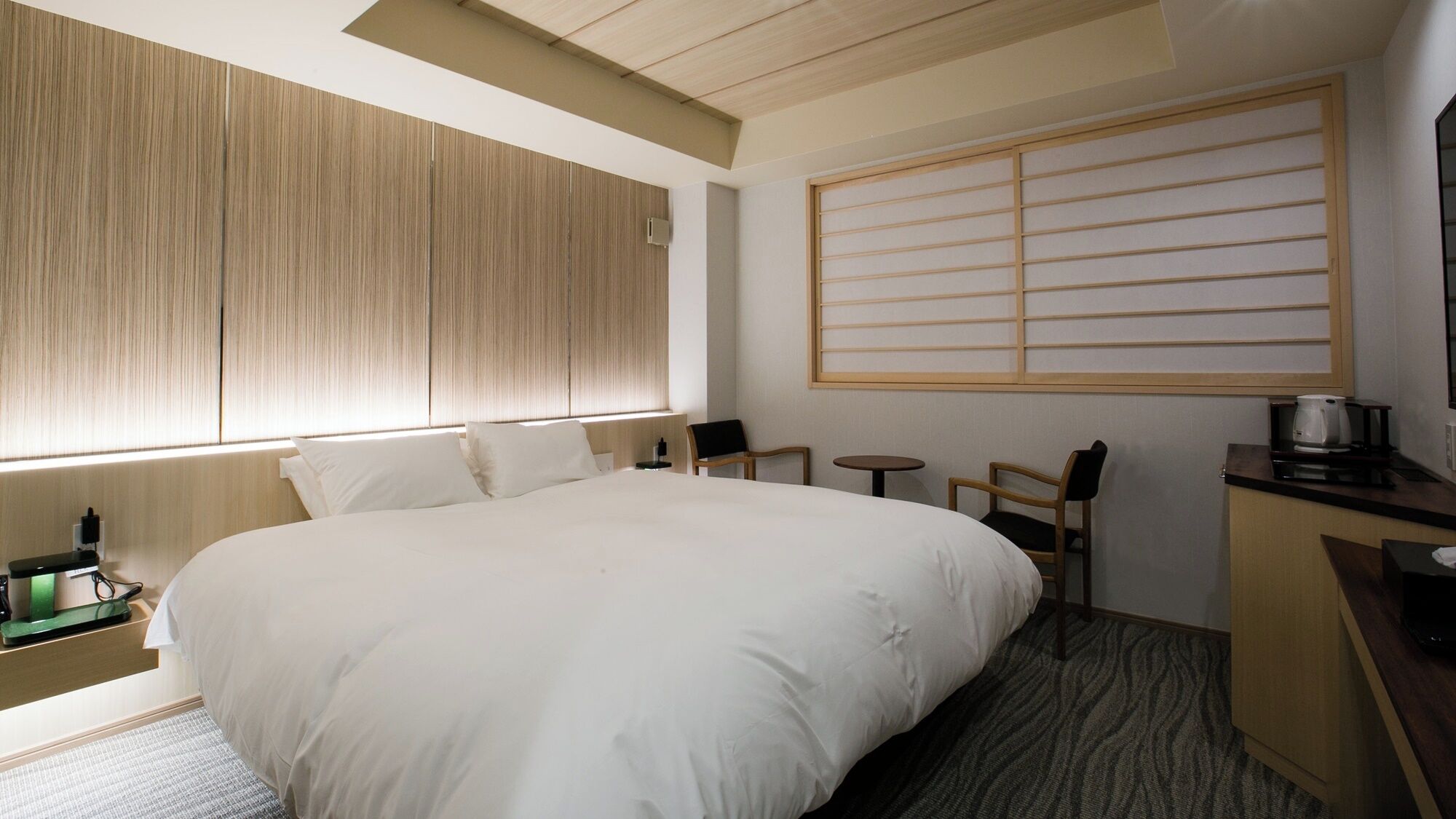 A hotel room with a large bed, a desk with a coffee maker, a small table, and a window with blinds.