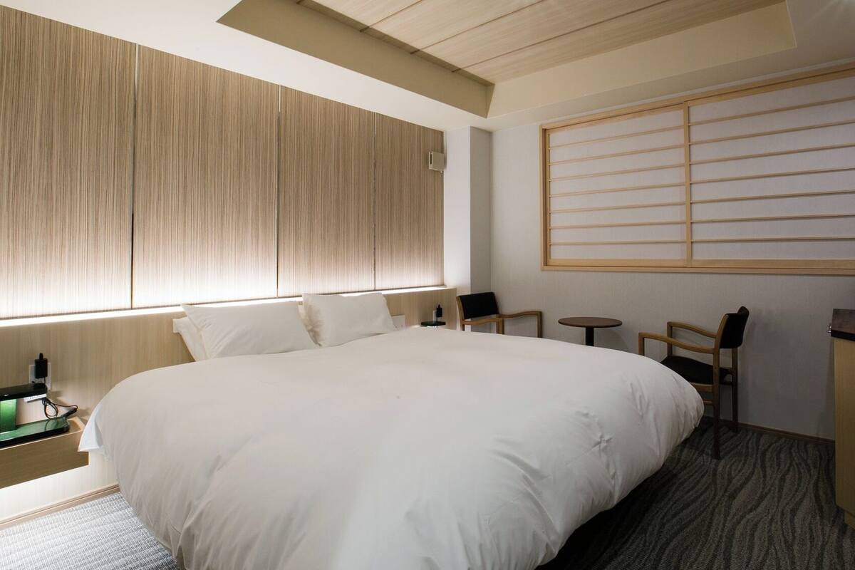 Habitación de hotel con una cama grande, un escritorio con cafetera, una mesita pequeña y una ventana con persianas.