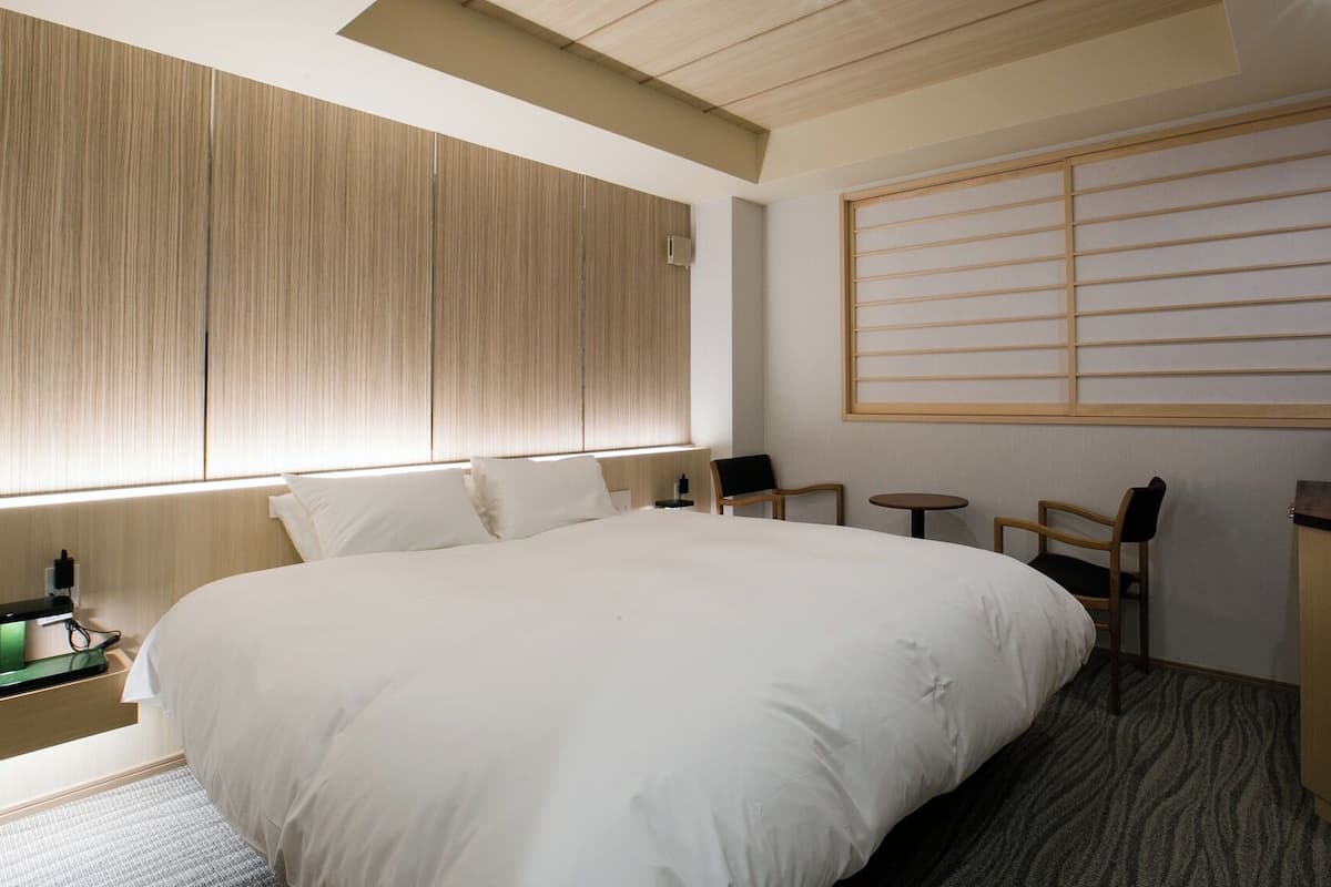 A hotel room with a large bed, a desk with a coffee maker, a small table, and a window with blinds.