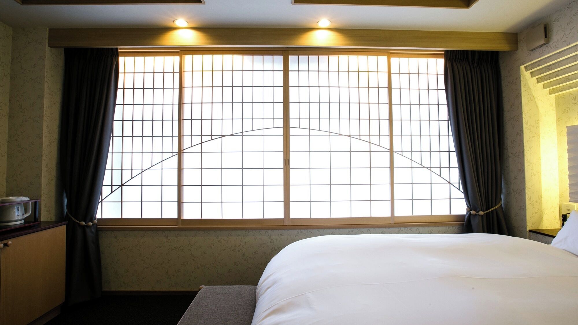 A hotel room with a bed, a nightstand, a window with curtains, and a wall-mounted heater.
