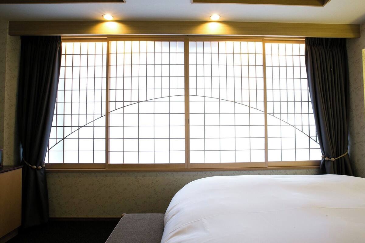 Una habitación de hotel con una cama, una mesita de noche, una ventana con cortinas y un calentador fijado a la pared.