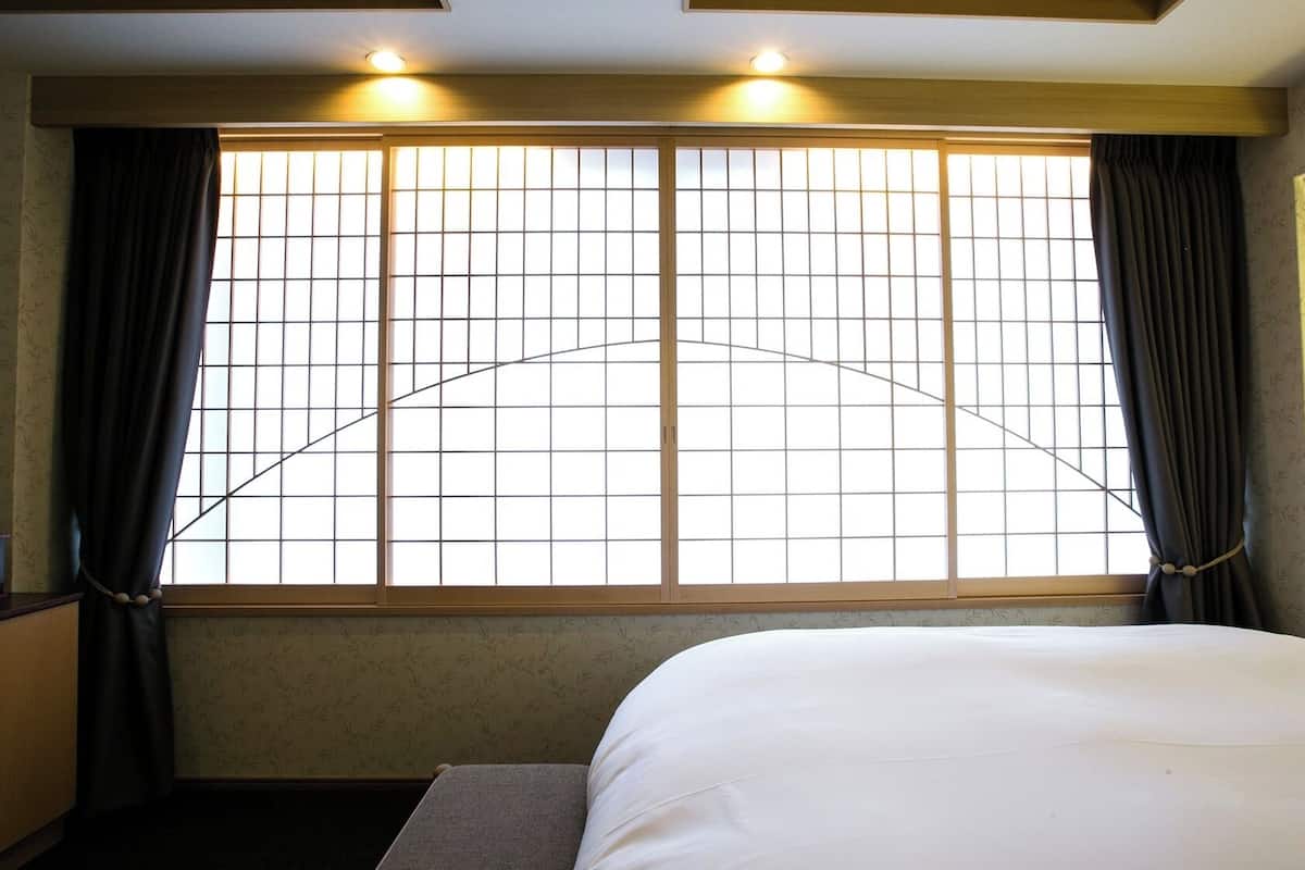 A hotel room with a bed, a nightstand, a window with curtains, and a wall-mounted heater.