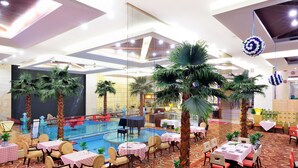 Vestíbulo de hotel con zona de piscina, varias mesas y sillas para comer, rodeado de palmeras.