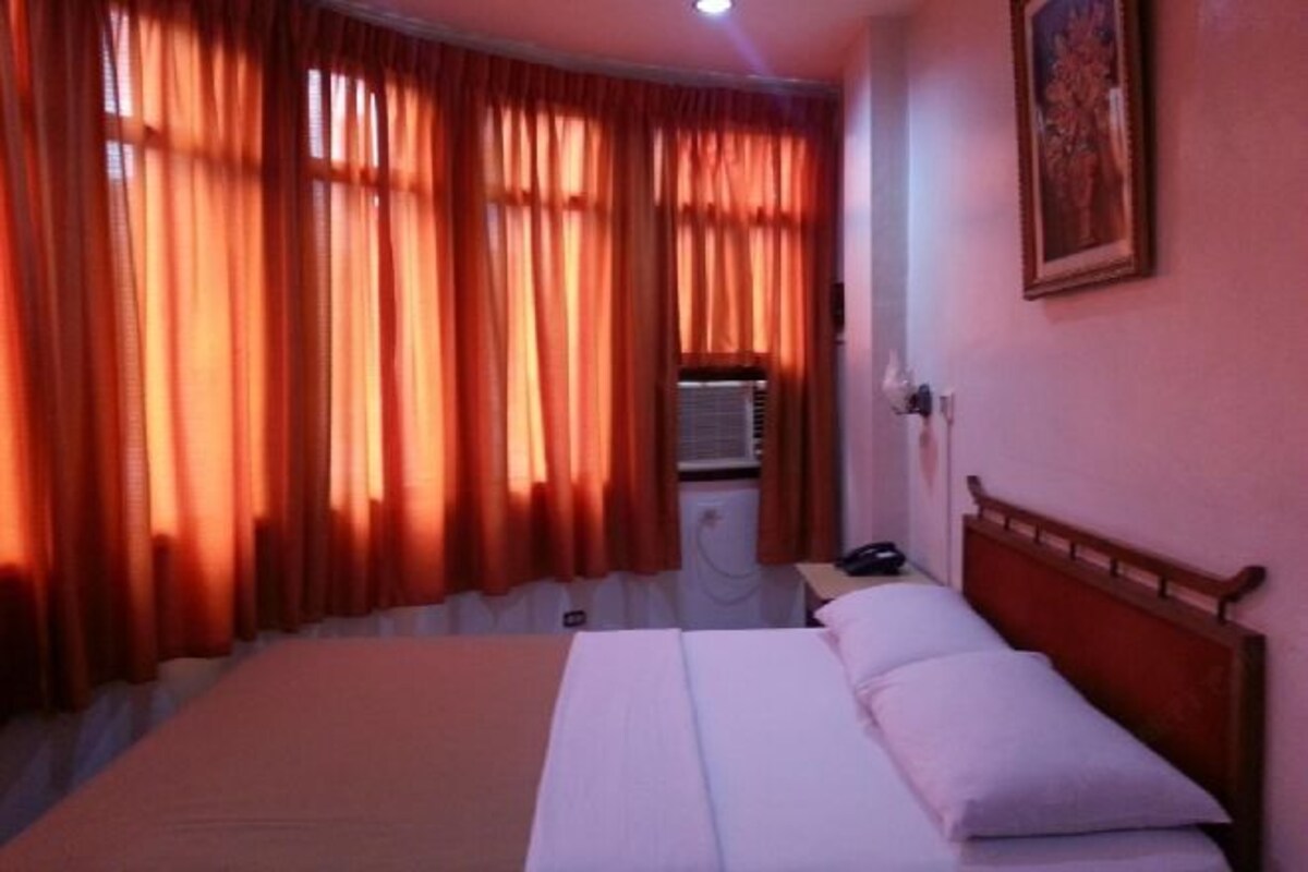 A hotel room with a bed, a nightstand, a wall-mounted phone, and a window with curtains.