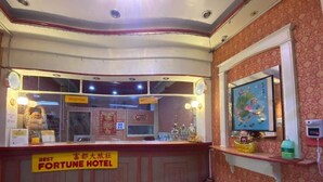 Un mostrador de recepción con un letrero que dice "BEST FORTUNE HOTEL" y un área de recepción con un mostrador, un extintor y una estantería con objetos decorativos.